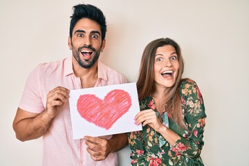 Beautiful young couple together holding heart draw celebrating crazy and amazed for success with open eyes screaming excited.
