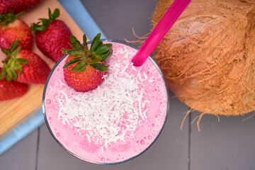 strawberry and coconut smoothie with a shredded coconut topping, a whole strawberry and pink straw. whole strawberries on a wooden board with a blue cloth napkin and a whole coconut on the side.