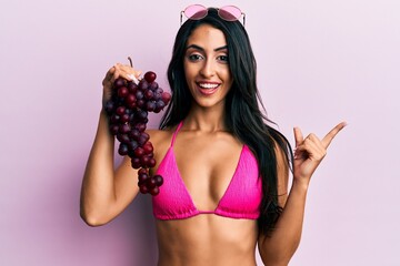 Beautiful hispanic woman holding branch of fresh grapes wearing bikini smiling happy pointing with hand and finger to the side