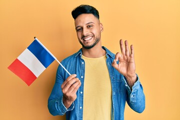Young arab man holding france flag doing ok sign with fingers, smiling friendly gesturing excellent symbol