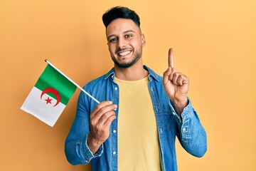 Young arab man holding algeria flag smiling with an idea or question pointing finger with happy face, number one