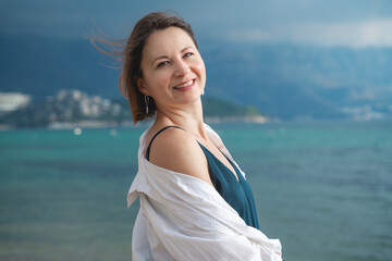 Cute smiling woman in white shirt poses on beach walking before storm, mountain on blurred background. Stay positive in the face of any adversity.