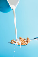 Pouring of milk from jug into spoon with cereal rings on color background