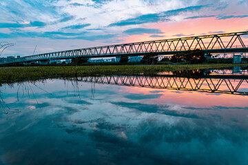 水溜りに反射する幾何学模様の橋と目が覚めるような夕焼けと河川敷