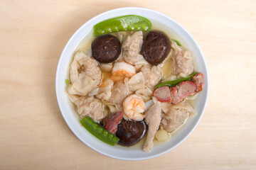 Close up view from above of a white porcelain bowl full of Wonton Soup on light brown table.