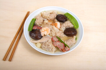 Close up view from above of a white porcelain bowl full of Wonton Soup with chop sticks laying on table. Light brown table.