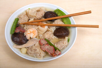 Close up view from above of a white porcelain bowl full of Wonton Soup with chop sticks laying on top. Light brown table.