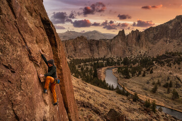Adventurous man is rock climbing on the side of a steep cliff. Sunset Sky Art Render. Taken in...