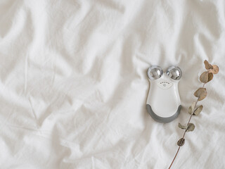 Microcurrent concept. Face massager with micro currents. Home use device for anti-aging and face lifting on white bed. Top view or flat lay, copy space for text.