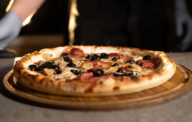 Hot pizza with olives and salami on a table on a wooden board in a pizzeria. Ready baked goods for...
