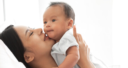 Portrait of enjoy happy love family asian mother playing with adorable little asian baby.Mom kiss with cute son moments good time in a white bedroom.Love of family concept