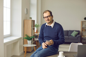 Portrait of successful businessman in glasses using smartphone sitting on edge of desk in his home office. Happy balding adult man in smart casual jumper and jeans text messaging on mobile phone
