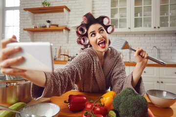 Food blogger, singer or social media addict having fun while cooking meal in kitchen. Young woman in hair curlers and housecoat singing in pretend spatula mic and taking funny selfie on mobile phone