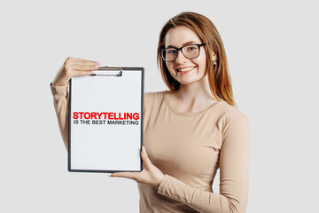 Storytelling is the best marketing. Beautiful young business woman wearing glasses holds a clipboard with mock up space isolated on gray background