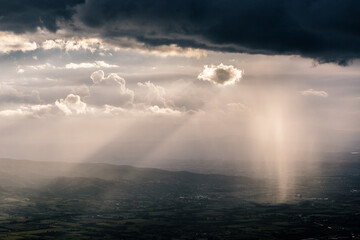 Rain, mist and sunrays between valley and layers of mountains and hills beneath a moody, overcast sky with heavy clouds