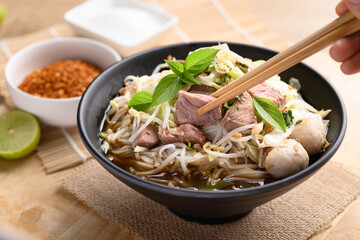 Hand holding chopsticks and eating rice noodle soup with pork, pork ball and vegetables in a bowl, Thai noodles soup