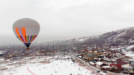 Aerial view of a hot air balloon over the icy  lake, snowy mountains around. hot air balloon in winter. Children are sledding. Glides over the camera in winter. People an ice slide. 
