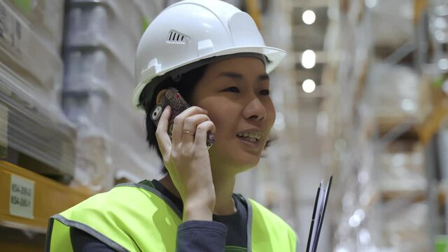 asian female warehouse worker with smartphone. holding clipboard. racks with supplies. concept logistics, merchandise, export. employee talking discussing on the phone