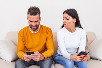 Jealous young woman with smartphone looking at smiling boyfriend using smartphone at home,...