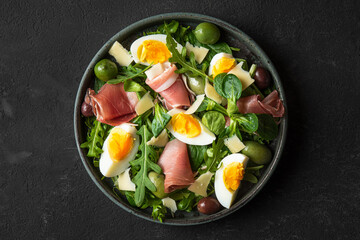 Fototapeta na wymiar Salad with prosciutto, olives, cheese, eggs and arugula in a plate on black background. Italian food snacks. Top view