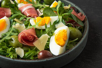 Salad with prosciutto, olives, cheese, eggs and arugula in a plate on black background. Italian food snacks. close up