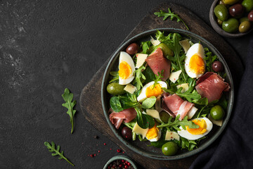 Prosciutto salad with parmesan, olives, eggs and argula in a plate on cutting board over black background. Italian food
