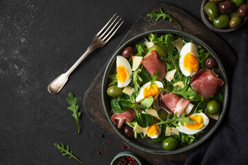 Healthy diet food. Salad with prosciutto, cheese, olives, eggs and arugula in a plate with fork on dark background. Top view