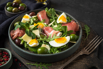 Prosciutto salad with parmesan cheese, olives, eggs and arugula in a plate with fork on cutting board over black background
