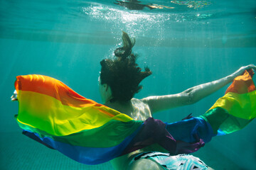 Horizontal view of unrecognizable woman swimming underwater holding a colorful lgbt flag. Lesbian,...