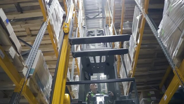Young man worker moving on loading machine during working day in modern warehouse. Front view of male employee is lowering pallet from shelves using equipment at industrial storehouse of factory