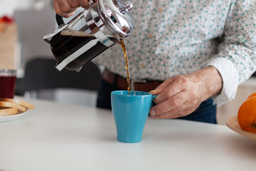 Senior man using french press and pouring hot drink after preparing coffee in kitchen for breakfast. Elderly person in the morning enjoying fresh brown cafe espresso cup caffeine from vintage mug