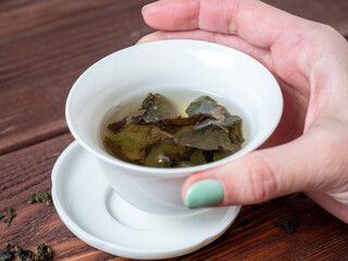 a woman's hand holds a white porcelain bowl with tea. Wooden background. The opened leaves of large-leaved tea. Chinese tea. tea party