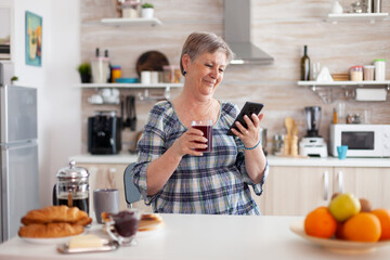 Senior woman using mobile gadget in the kitchen drinking aromatic tea in the morning during breakfast. Authentic elderly person searching on modern smartphone internet technology, senior leisure time - Powered by Adobe