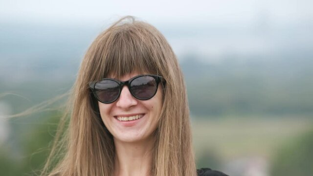 Portrait of a happy young long haired woman in dark sunglasses smiling in camera on a windy summer day.