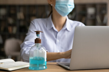 Crop close up of bottle with liquid antibacterial sanitizer on table in office to protect from coronavirus infection. Woman work online on computer wear facial mask from covid-19. Corona concept.