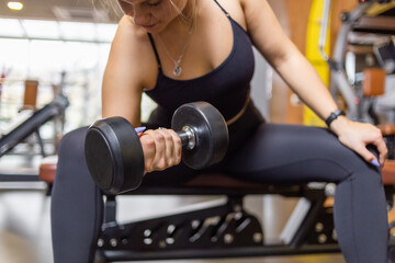 Fototapeta na wymiar A woman trains with a dumbbell in her hand while sitting on a bench in the gym. Concentrated biceps dumbbell curls