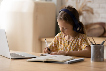 Good pupil. Little girl get remote distant education online listen to teacher tutor at virtual class lesson take write notes by hand. Schoolchild in headphones use modern tech in learning by internet