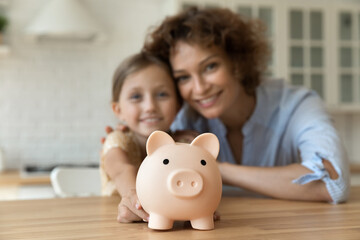 Take care of future. Blurred portrait of smiling young mother small girl daughter look at camera show funny pig for coins offer you to make bank deposit save money. Focus on piggybank on kitchen table