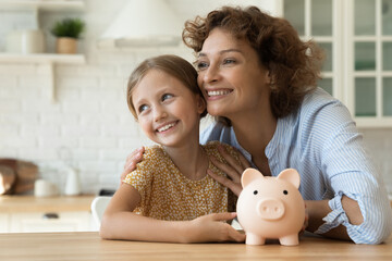 Loving family of young mother little daughter hug by kitchen table hold cute piggybank look away...