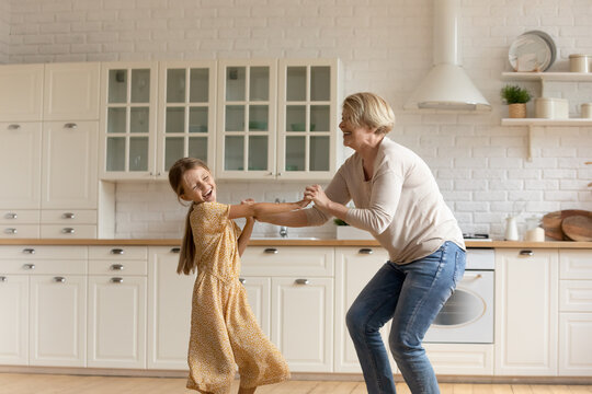 Granny teaches to dance. Laughing little girl mature aged grandmother hold hands move by energetic music having fun. Active senior grandma entertaining small grandchild kid sing jump at home kitchen