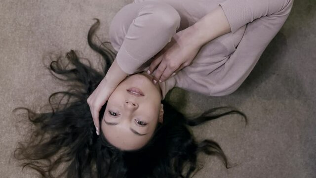 A happy, smiling, dark-haired woman lying on the floor is enjoying a rest in a bright room. Top view.