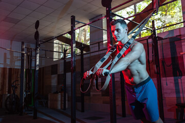 Muscular man works out with the trx system in a dark gym. Athlete with naked torso workout with resistance straps in red-blue neon light. Functional, cross training