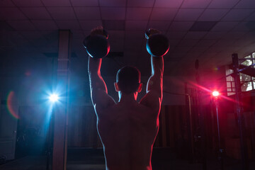 Muscular Caucasian man with naked torso training overhead with kettlebells in red blue neon...