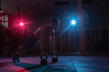 Muscular man push up with kettlebell in red-blue gradient light in dark cross fit hall