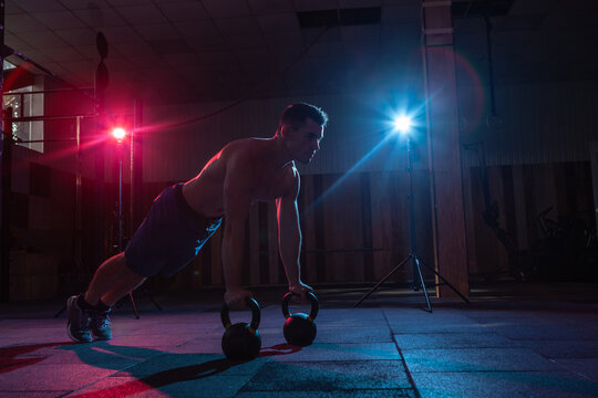 Muscular man push up with kettlebell in red-blue gradient light in dark cross fit hall