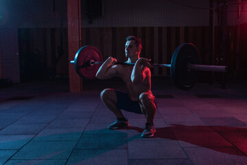 Obraz na płótnie Canvas Working out by lifting weights in a cross-training gym. Muscular powerful man with heavy barbell doing squat exercise in red blue neon light