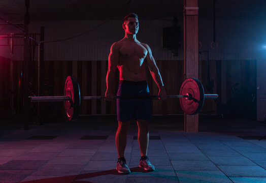 Working Out By Lifting Weights In A Cross-training Gym. Muscular Powerful Man Doing Deadlift With A Heavy Barbell In Red Blue Neon Light