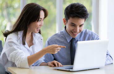 Beautiful black long hair Asian businesswoman and gook looking cute Asian businessman sitting together and watching the screen of laptop notebook computer in office with smile face