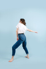 Lively contemporary dancer poses in front of blue studio background. She's lightly arching her back, legs placed solidly, making twisting motion with outstretched hand.