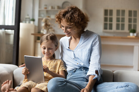 Young female babysitter look after little child girl sit on couch help study language on digital tablet using web app for children. Interested mother hug small kid daughter read ebook from pad screen